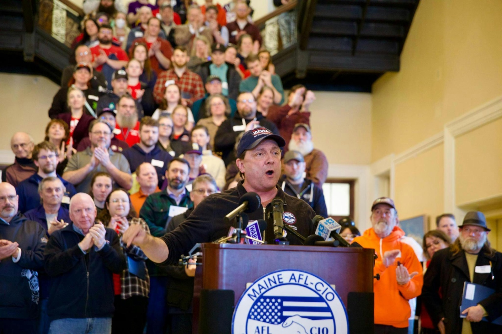 Troy Jackson speaking at the Maine AFL-CIO podium with a crowd of union members behind him