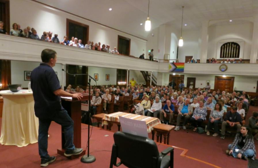 Troy Jackson speaking at a packed town hall in Gorham, Maine, with an empty chair onstage
