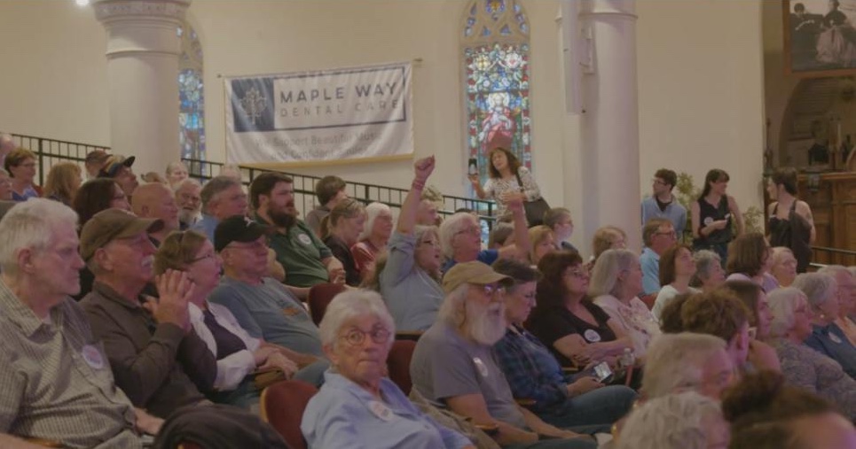 A packed hall in Lewiston, Maine, with a voter raising a hand to ask a question