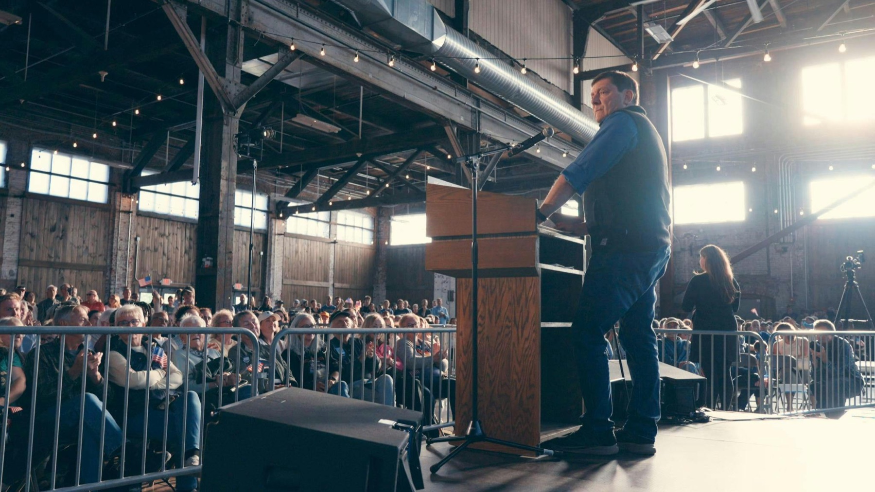 Troy Jackson at a podium in front of a packed crowd in a brick hall